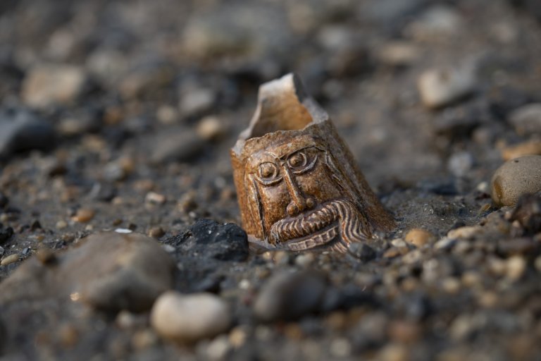 A carved broken pipe bowl shaped like a bearded face lies half buried among pebbles on the ground © Alessio Checconi London Museum 1 768x512